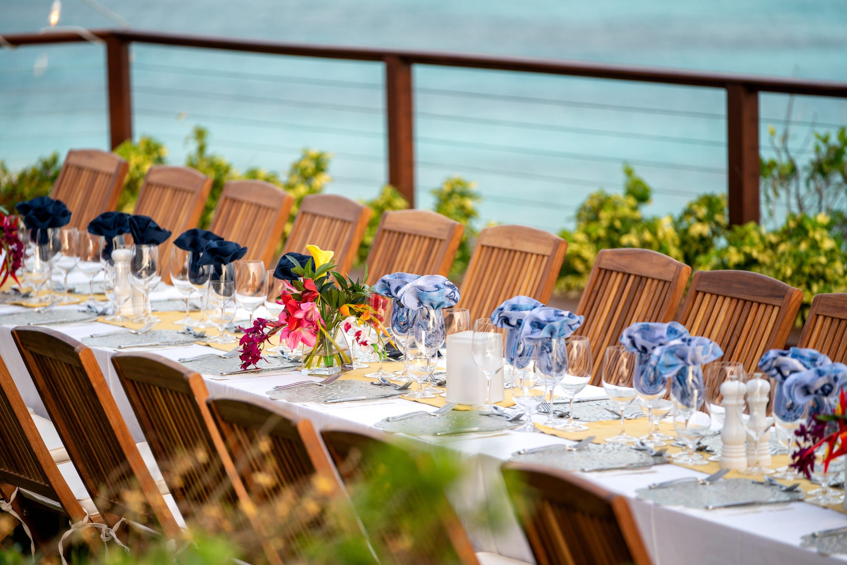 A long table laid for dinner on a terrace above the sea, with folded napkins, flowers and glassware